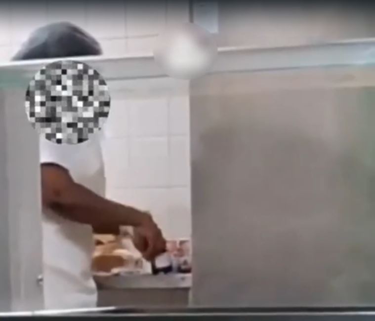 No registro, a colaboradora aparece utilizando os próprios dedos para espalhar geleia em fatias de pão dentro da área de preparação das refeições